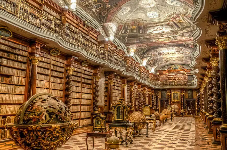 intérieur de la bibliothèque du Clementinum à Prague. Le sol carrelé et le plafond peint sont ouvragés. Des meubles et des sculptures précieux ornent la galerie. Des milliers de livres garnissent les murs du sol au plafond.