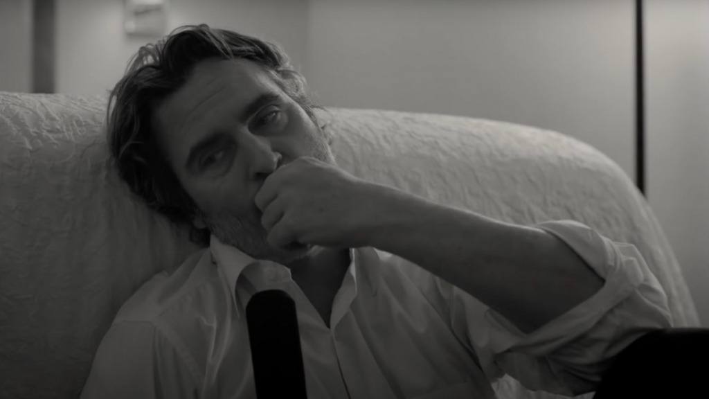 Image en noir et blanc de Joaquin Phoenix dans un de ses rôles. Un homme d'une cinquantaine d'année assis sur un canapé, le regard trist et dans le vague, le point porté à la bouche.
